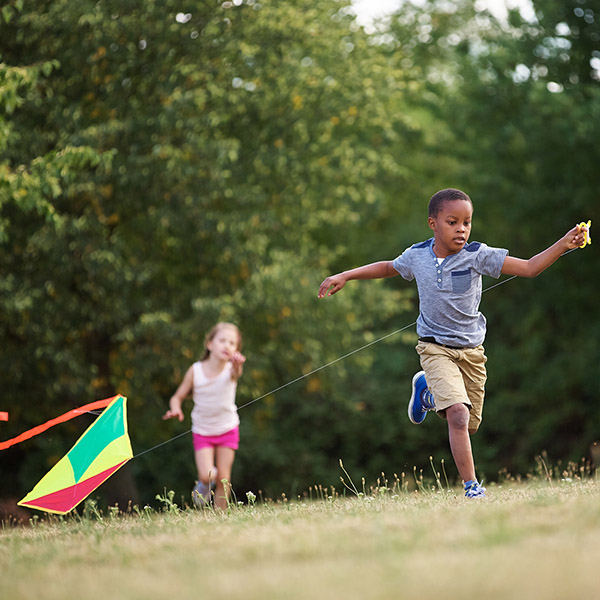 Two children run through a park with a kite following behind them.